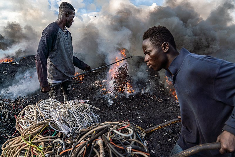 A young man is pictured burning electrical wires to recover copper at Agbogbloshie, Ghana, as another metal scrap worker arrives with more wires to be burned. (Image by Muntaka Chasant, published under a <a href="https://spdx.org/licenses/CC-BY-SA-4.0.html">CC-BY-SA-4.0</a> license.)